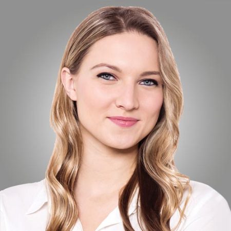 A young woman with long, wavy blonde hair and blue eyes, wearing a white blouse, smiles softly at the camera against a neutral gray background.