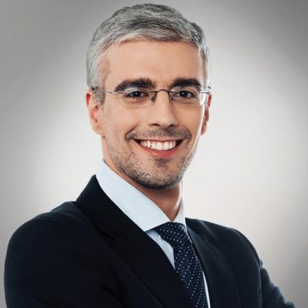 A man with short gray hair, wearing glasses, a dark suit, a light blue shirt, and a navy tie with small white dots, is smiling confidently at the camera against a neutral background.